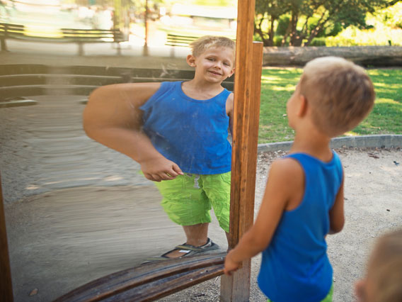A boy looking at a mirror where the perspective makes him look like his arm is much larger than it actually is.